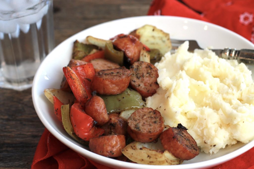 cooked sausage with red and green bell peppers, with a side of mashed potatoes, served in a white bowel with a glass of ice water.