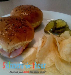 Baked ham and cheese slider served with a side of pickles and potato chips all on a white plate.