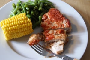 Slow cooked pork chops topped with chili sauce, served with a side of green beans a petite ear of corn, all served on a white plate.