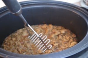 Cooked meat in a pressure cooker being mashed by a mashed potatoes masher.