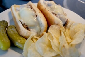 two sloppy philly cheese steak, on white hotdog buns, served with a side of potato chips and two dill pickles on a white plate.