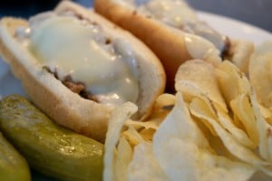 two sloppy philly cheese steak, on white hotdog buns, served with a side of potato chips and two dill pickles on a white plate.