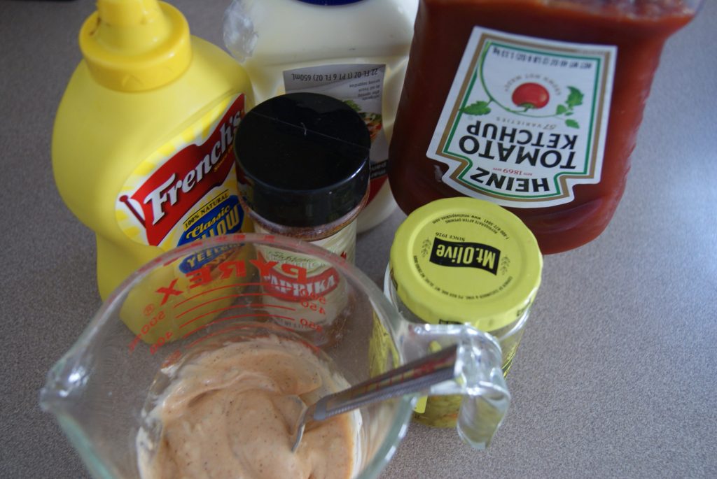 Bottles of yellow mustard, ketchup, paprika, relish, mayonaise, mixed up in a mixing cup for the secret sauce.