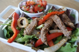 Seasoned beef fajitas, served with red bell peppers and onions on top of a salad, with a side of pico.