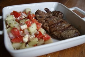 Greek grilled steak in a white dish served with a side of cucumber and petite tomato salad.