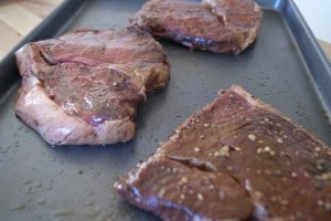 Three pieces of steak, seasoned and placed on a baking sheet.