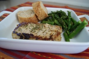 One pork chop seasoned with herbs and served on a white plate with a side of asparagus and two slices of bread.