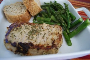 One pork chop seasoned with herbs and served on a white plate with a side of asparagus and two slices of bread.