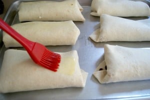 Wrapped tortillas being drizzled with melted butter on a baking sheet ready to be heated.