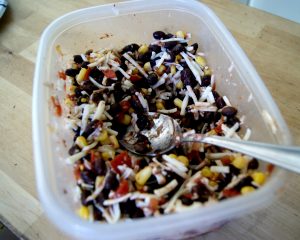 Black beans, shredded cheese, corn, salsa, and cumin all mixed in a plastic container.