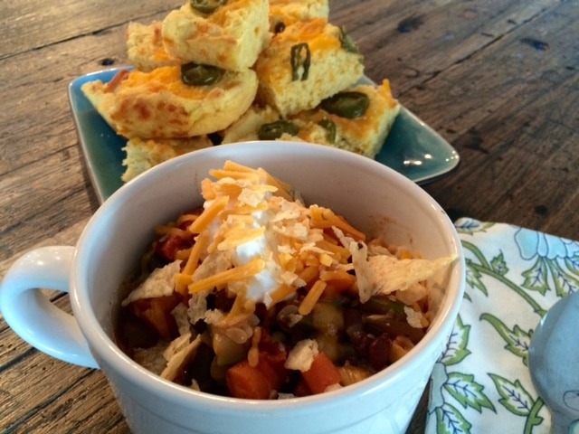 Slow cooked vegetable chili, in a white bowl, topped with sour cream and shredded cheese, with a side of jelepano bread.