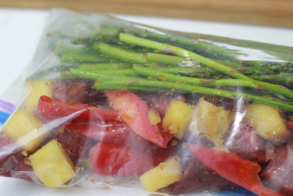 Paleo ginger steak, in a clear plastic bag with pineapple, asparagus, and sliced red bell peppers.