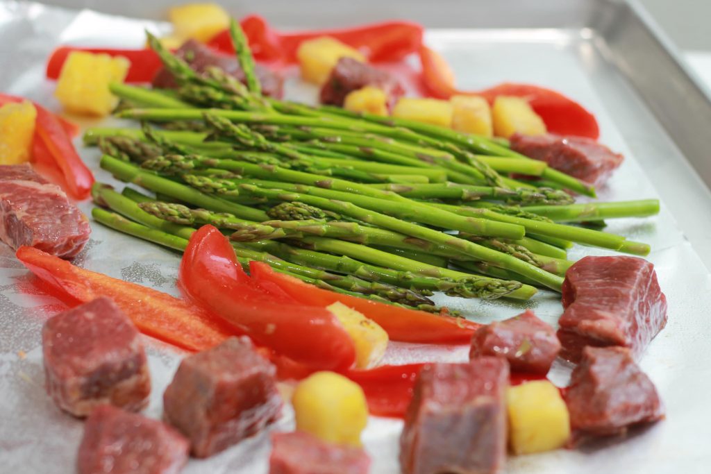 Asparagus, chopped pineapples, sliced red bell peppers, and steak all placed on one pan ready to be cooked.