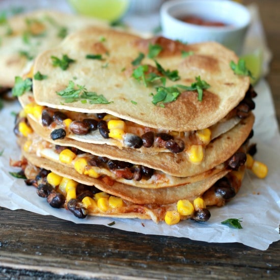 Three corn, black bean, salsa, and shredded cheese quesadillas stacked on top of one another served with a side of salsa and a lime, toped with seasoning.