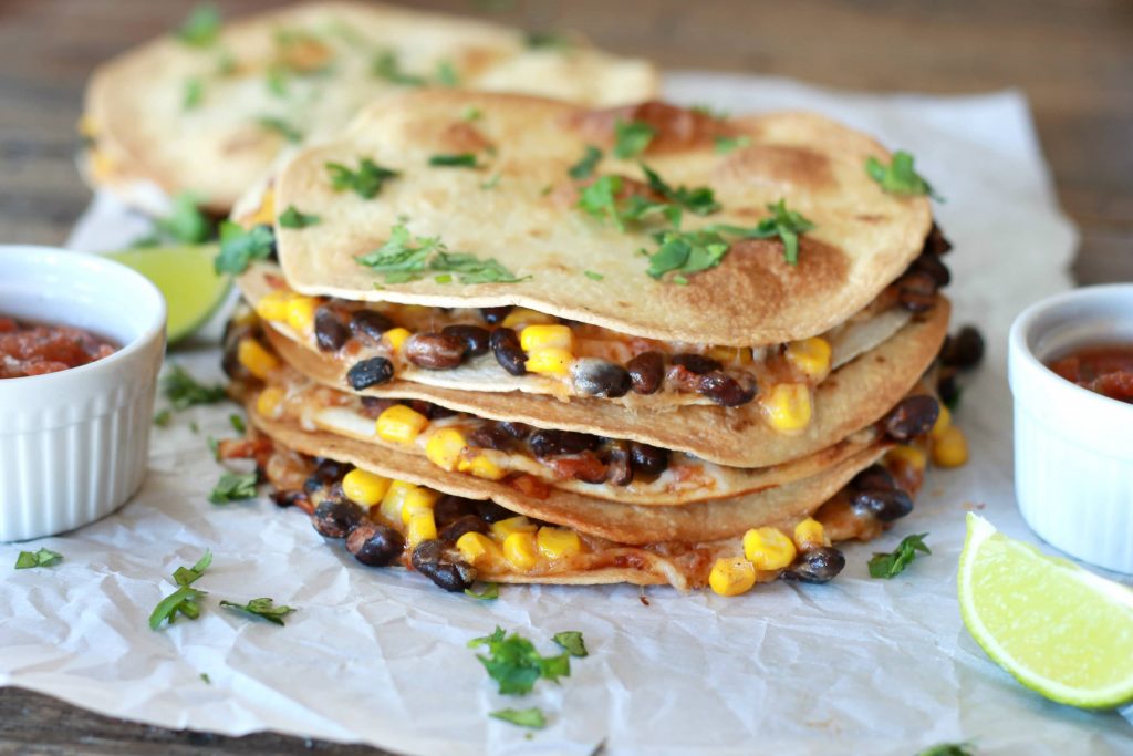 Three corn, black bean, salsa, and shredded cheese quesadillas stacked on top of one another served with a side of salsa and a lime, toed with seasoning.