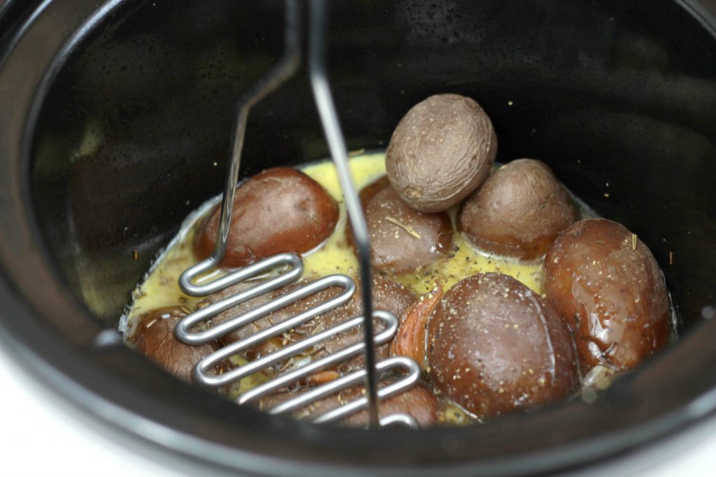 Cooked petite red potatoes getting mashed by a masher in the crockpot while sitting in melted butter, and milk.
