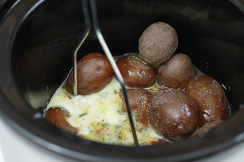 Cooked petite red potatoes getting mashed by a masher in the crockpot while sitting in melted butter, and milk.