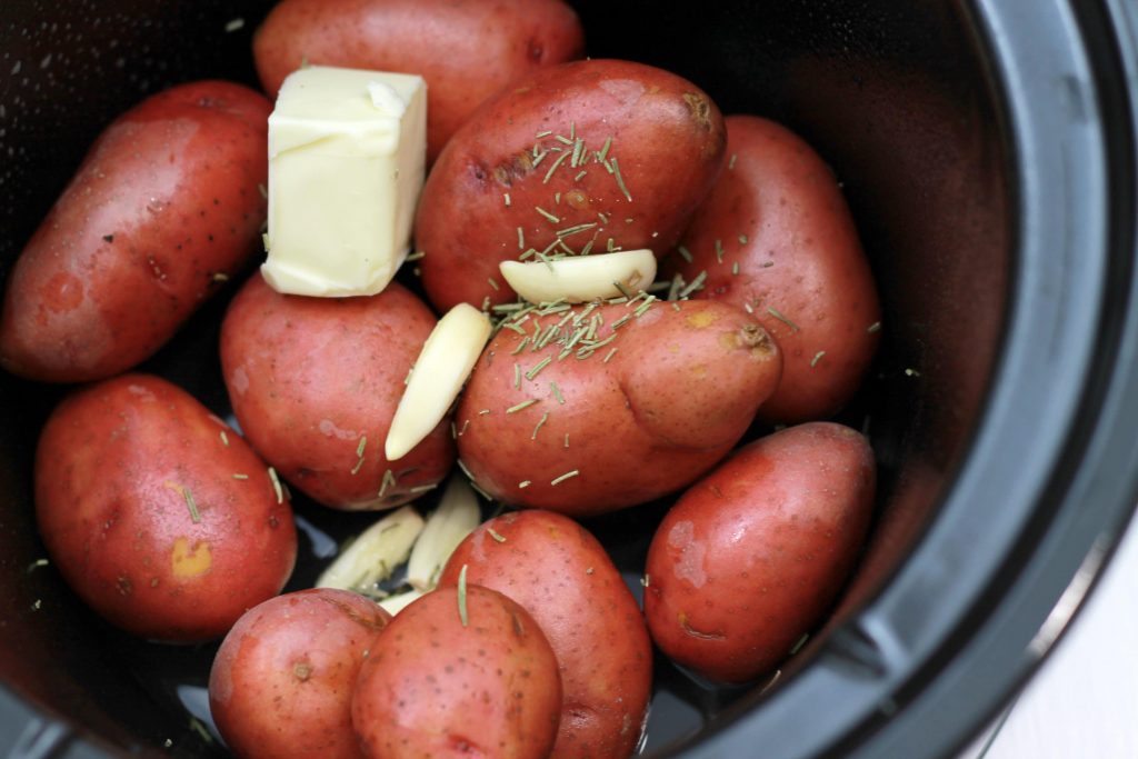 Unpeeled petite red potatoes placed in a crock pot topped with butter, clovers of garlic and seasoning. 