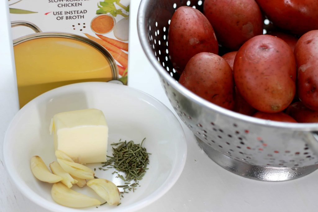 A box of chicken broth, a bowl with a fourth stick of butter, garlic cloves, and seasoning, next to a colander filled with washed petite red potatoes.