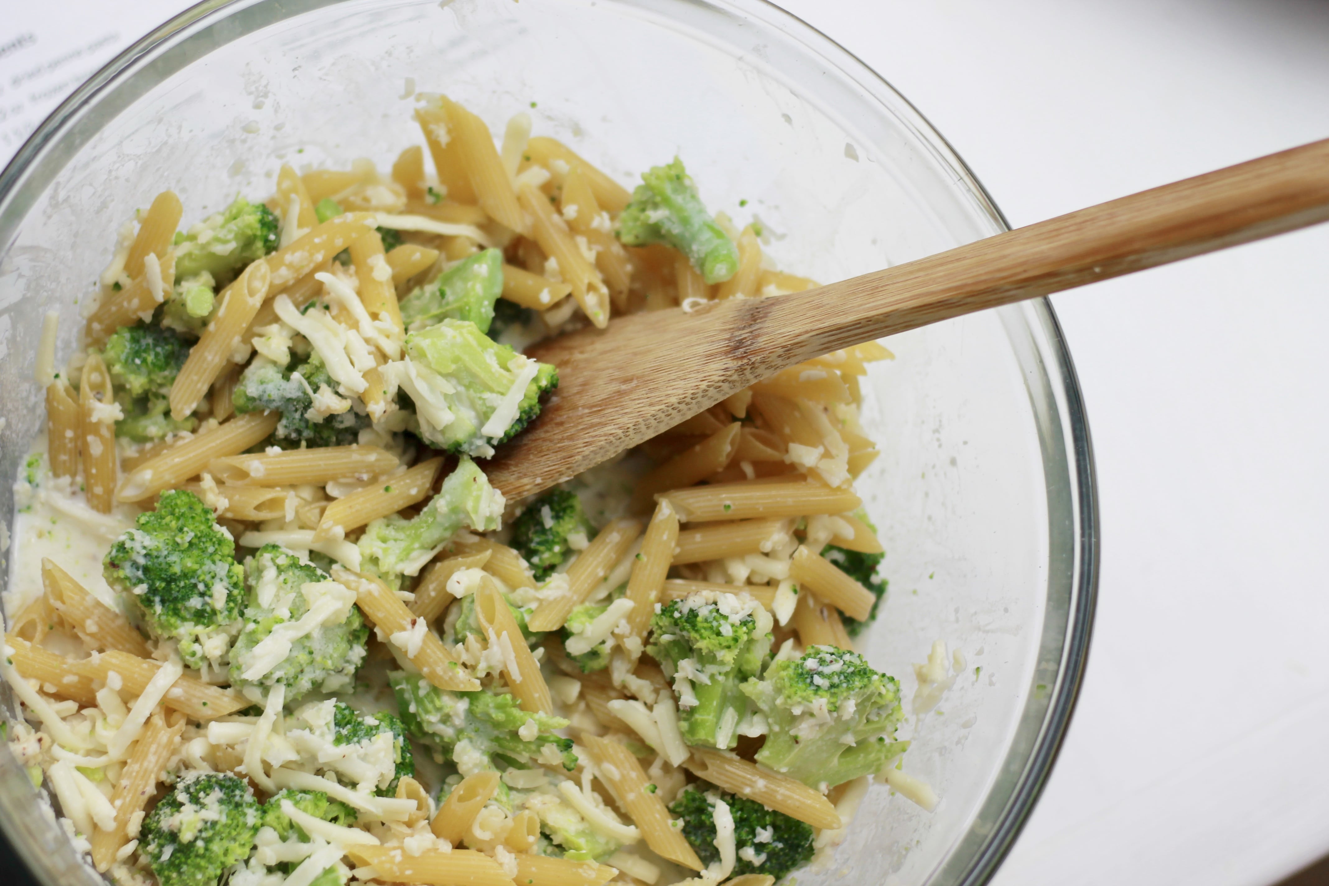 Broccoli, noodles and shredded cheese all mixed together in a clear bowl.