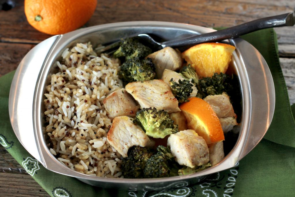 Orange chicken, with broccoli, and cooked diced oranges served with a side of brown rice in a silver dish ready to be eaten.