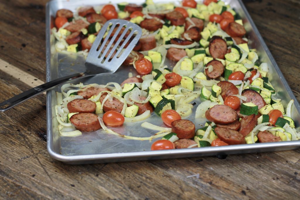 Sausage, zucchini, petite tomatoes, and chopped onions all cooked in one pan being served by a spatula.