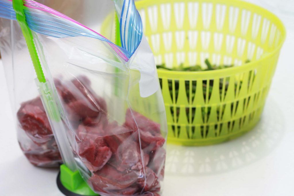 uncooked steak bites in a plastic bag, held up by green hands free baggie holder, with rinsed asparagusin a green basket.