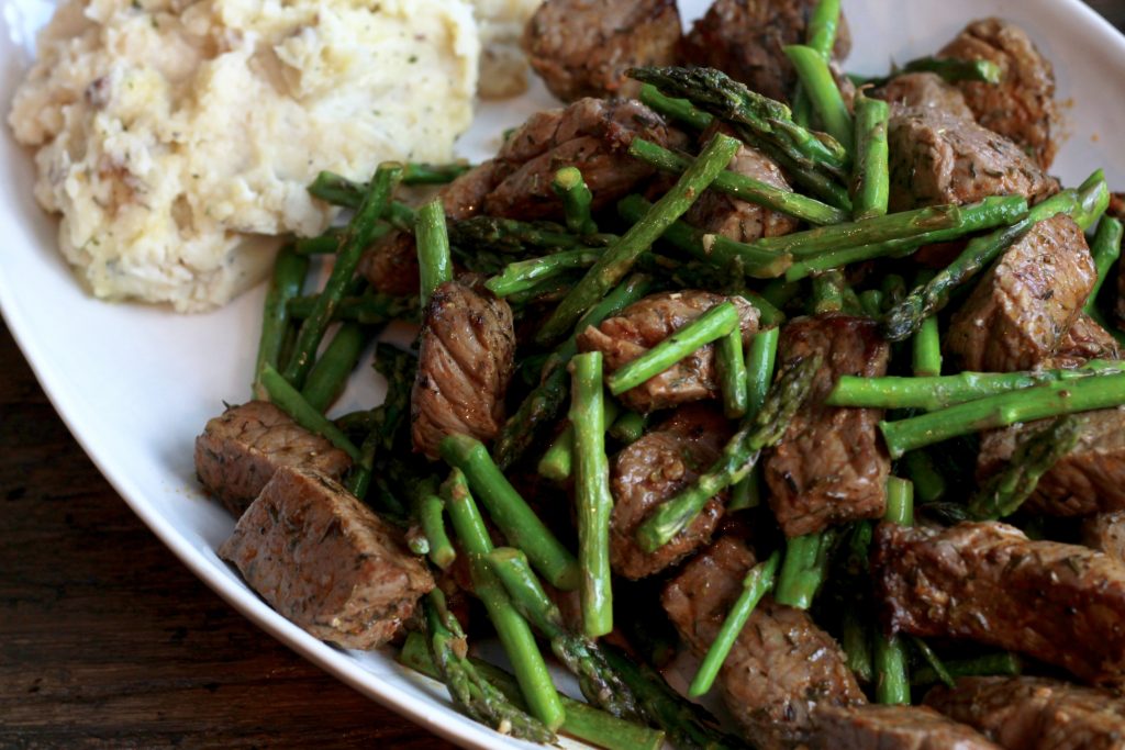 spices steak bites with asparagus with a side of mashed potatoes on a white oval plate.