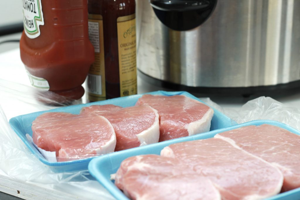 six uncooked slices of pork ready to be rinsed and cooked next to a bottle of ketchup and BBQ sauce.