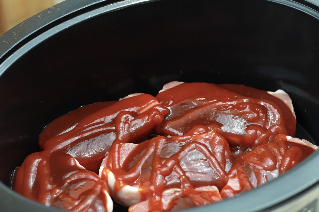 Six pork slices placed in a crockpot topped with BBQ sauce and ketchup ready to cook.