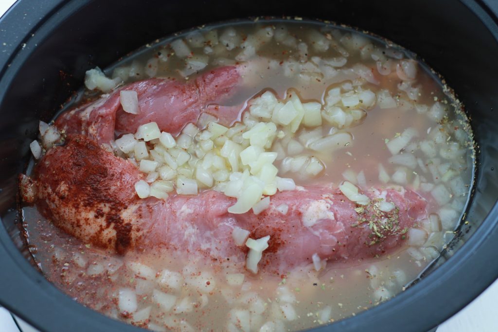 Large pork tenderloin soaking in chicken broth, chopped onions, and seasoning ready to sit and cook in the crockpot.