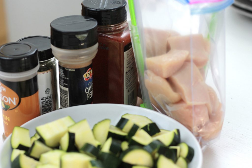 Chopped cucumber in a white bowl, with a clear plastic bag full of diced chicken, with four spices next to it. 