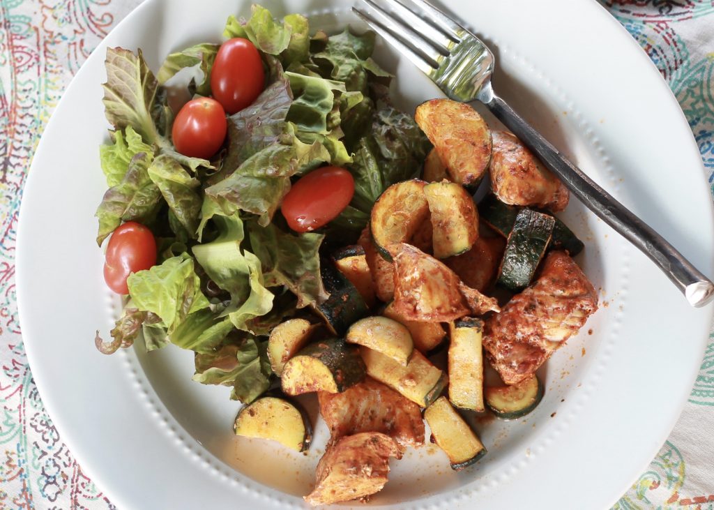 Cooked marinated chicken, and cucumbers on a white plate served with a side salad with petite red tomatoes ready to be eaten.