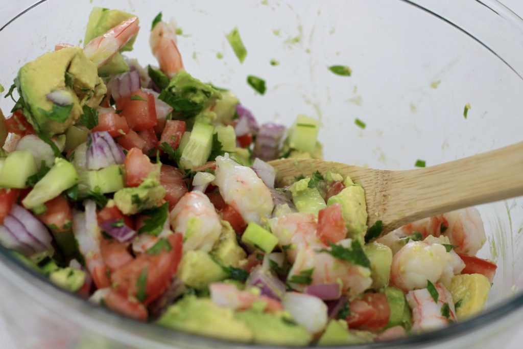 Shrimp, avocado, chopped onions, tomatoes, chives, and cilantro, all being mixed together in a clear bowl.