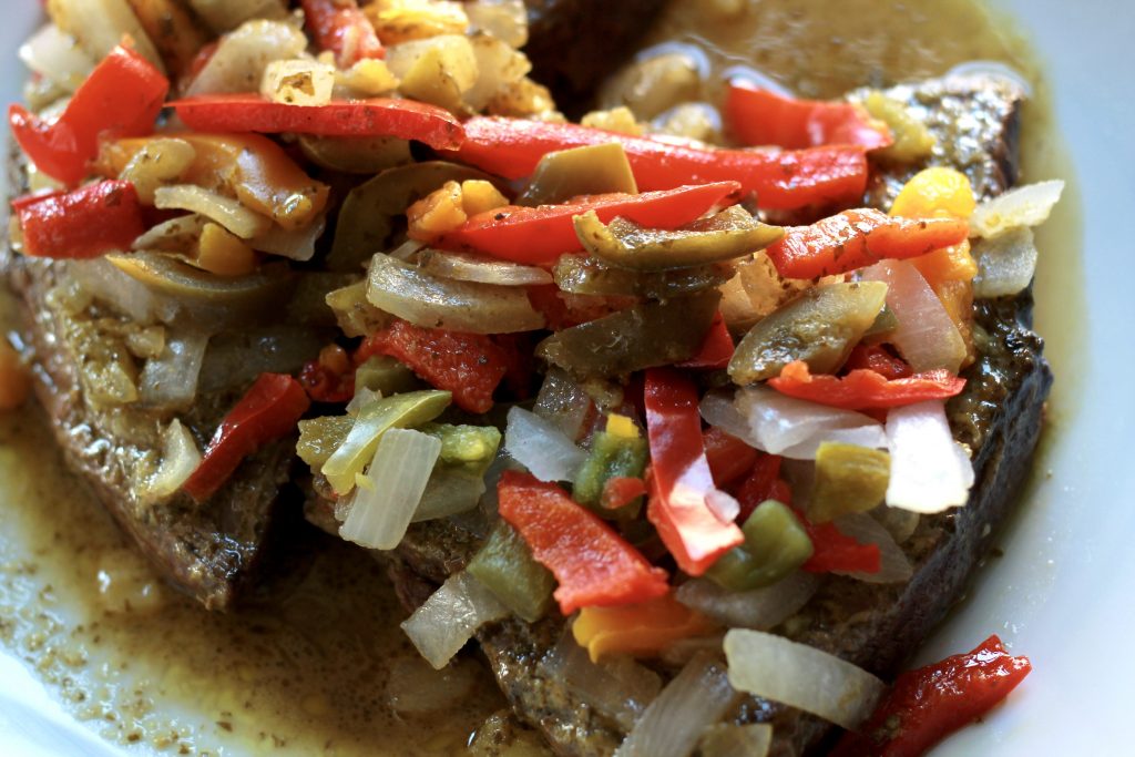 Cooked flank steak topped with onions, bell peppers, and vegetables, served on a white plate.