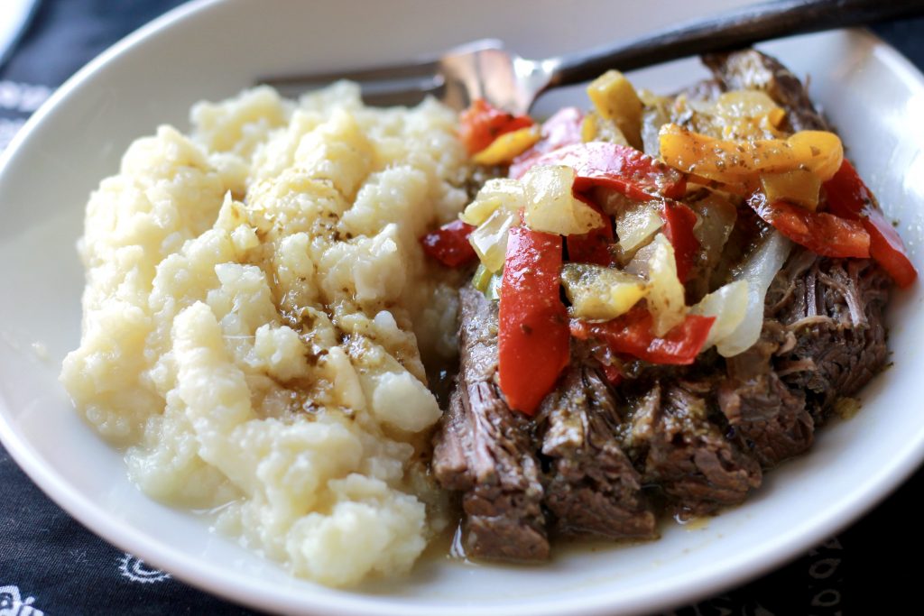 Cooked flank steak topped with onions, bell peppers, and vegetables, served on a white plate with a side of cauliflower mashed potatoes.