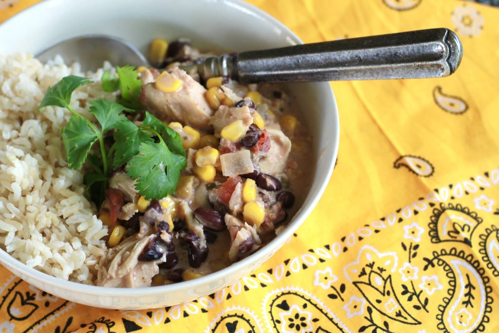 Cooked diced chicken mixed with corn, onions, tomatoes and black beans served with a side of white rice topped with cilantro all in white bowl .