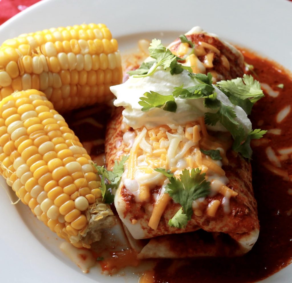 Slow cooked beef burrito wrapped in a four tortilla, bathed in enchilada sauce, topped with shredded cheese an lettuce, with a side of two petite ears of corn all served on a white plate.