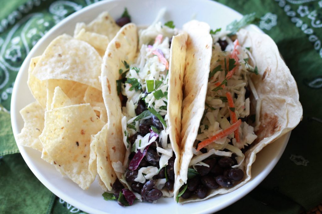 Two flour tortilla soft tacos with beans and cilantro slaw served with a side of tortilla chips on a white plate.