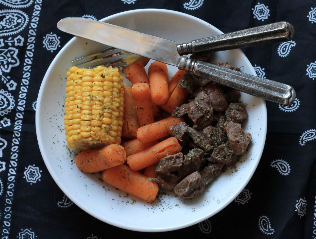 Cooked seasoned chopped steak, served with a side of slow cooked carrots and a petite ear of corn,all served on a plate.
