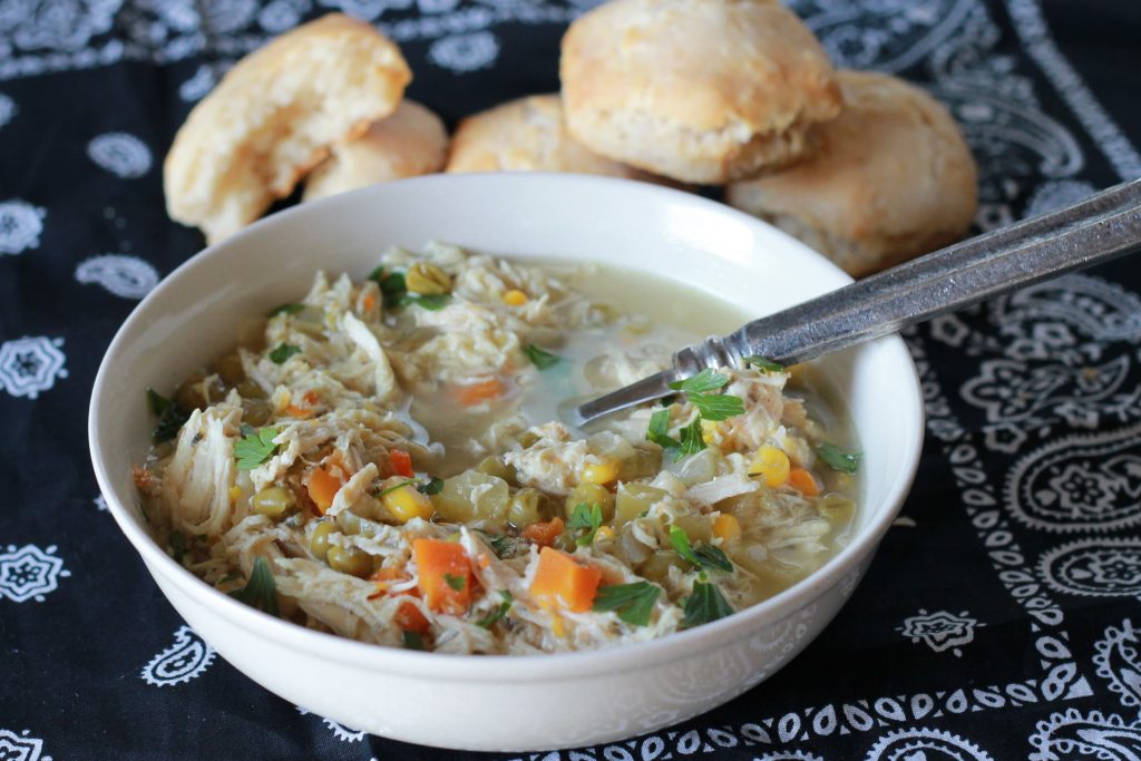 slow cooked chicken with chunks of carrots, corn and broth in a white bowl served with a side of biscuits, on a blue bandana.