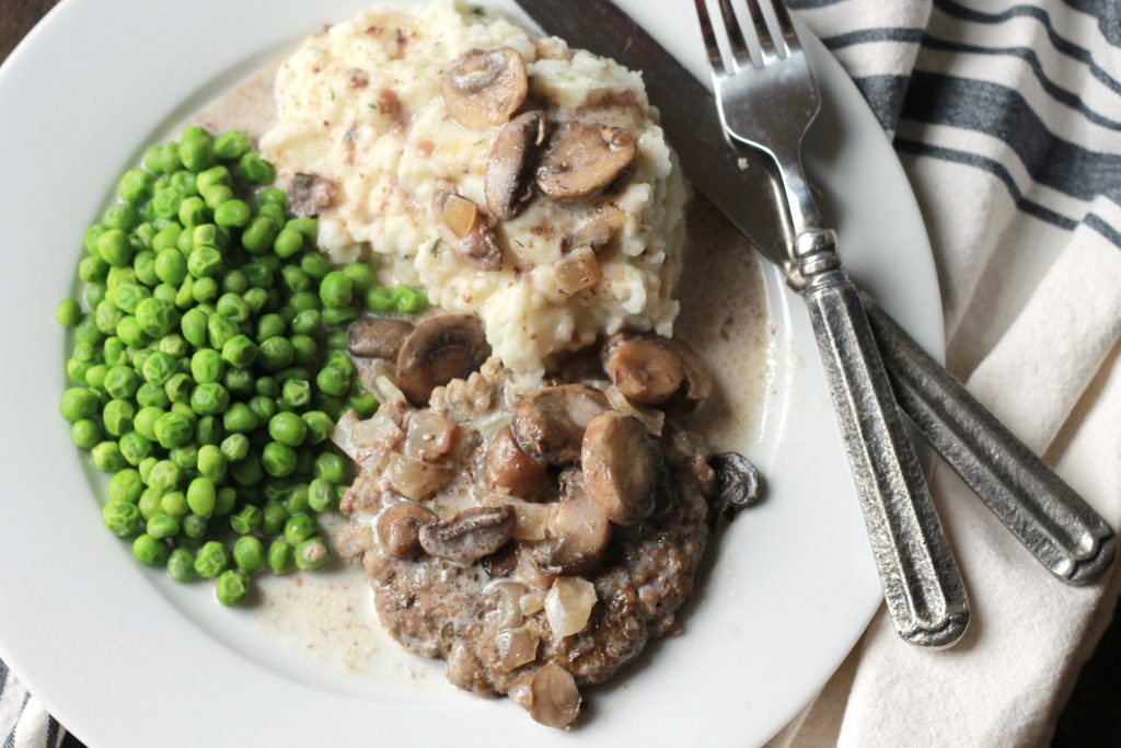Slow Cook Salisbury Steak in Cream Gravy with a side of green peas, and mashed potatoes served on a white plate with a silver fork and knife