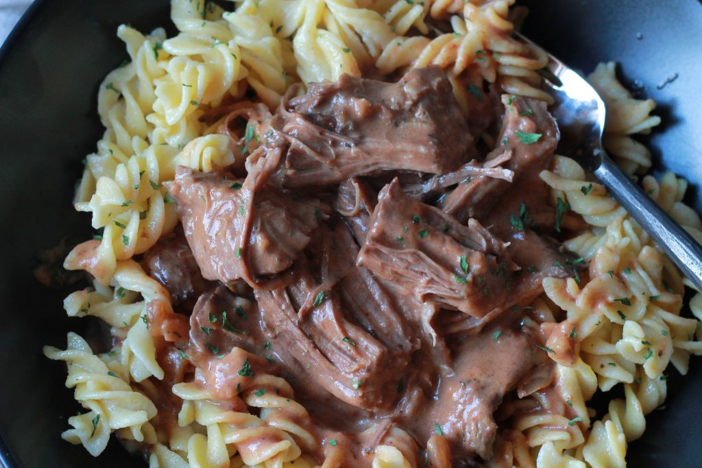 slow cooked beef with creamy tomato sauce seasoned sauce served on top of cooked pasta in a black bowl.
