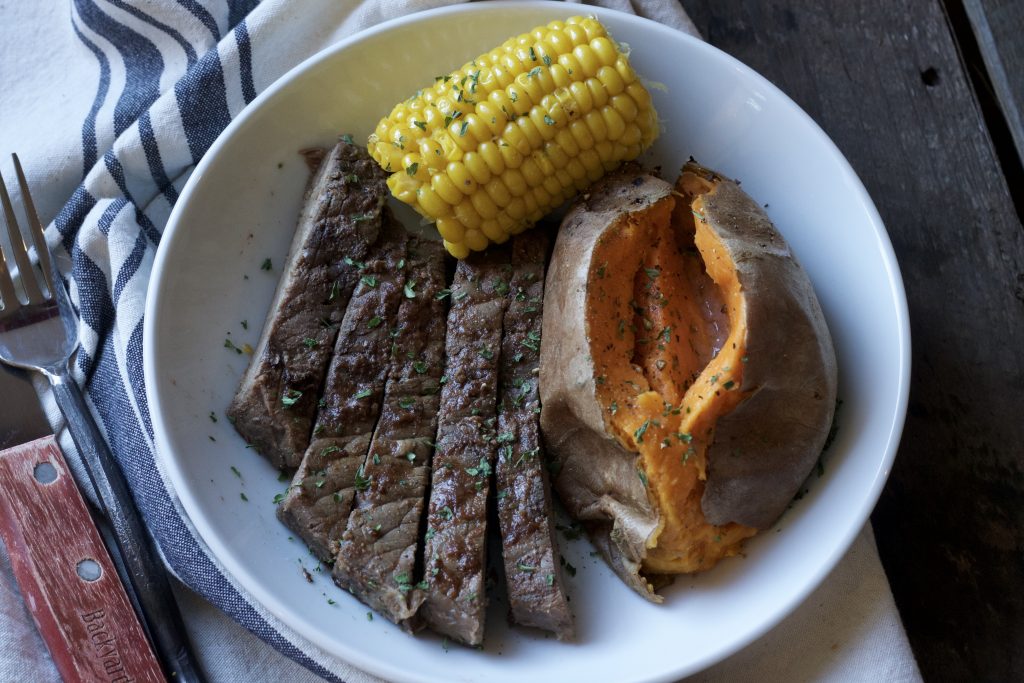 Tender seasoned steak served with a petite ear of corn and one cooked sweet potato on a white plate. With a side of biscuits on a white and blue napkin. 