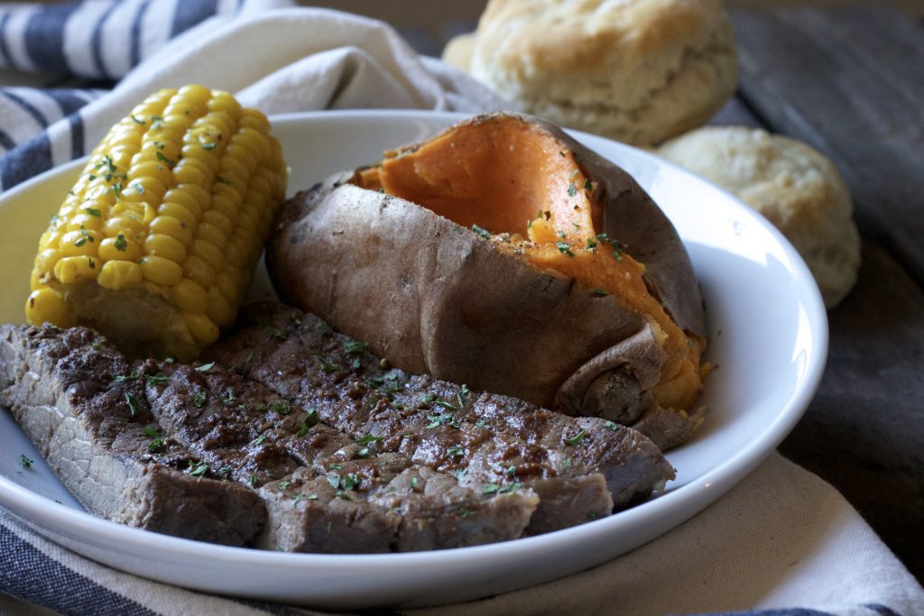 Tender seasoned steak served with a petite ear of corn and one cooked sweet potato on a white plate. With a side of biscuits on a white and blue napkin. 