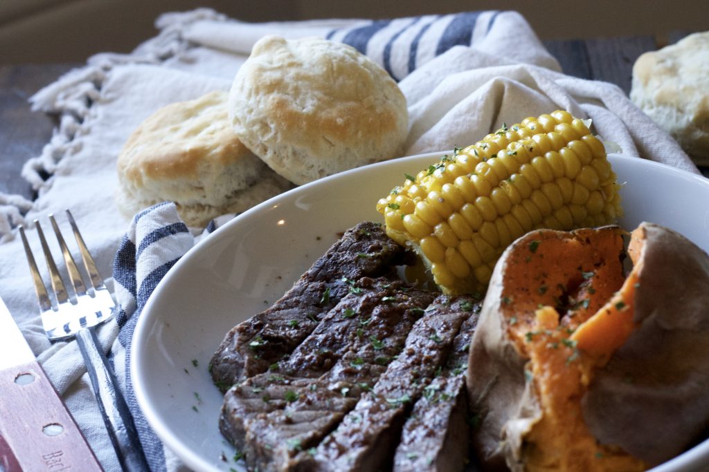 Tender seasoned steak served with a petite ear of corn and one cooked sweet potato on a white plate. With a side of biscuits on a white and blue napkin. 