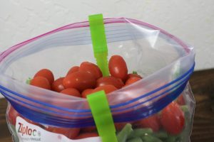 one green Jokari Hands-Free Bag Holder with a clear zip lock bag filled with petit red tomatoes and frozen green beans.