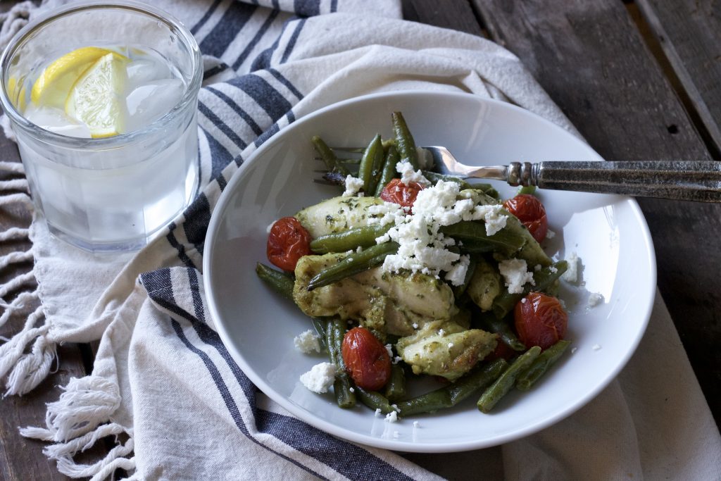 mixed chicken with green beans, petite tomatoes all dressed in basil pesto sauce served in a white bowl with a silver fork topped with parmesan cheese with a glass of lemon water