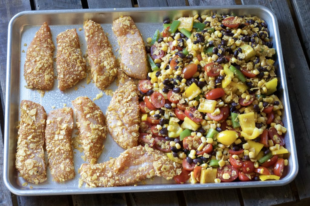 Chicken strips covered with tortilla strips on a sheet pan next to black bean and corn mix.