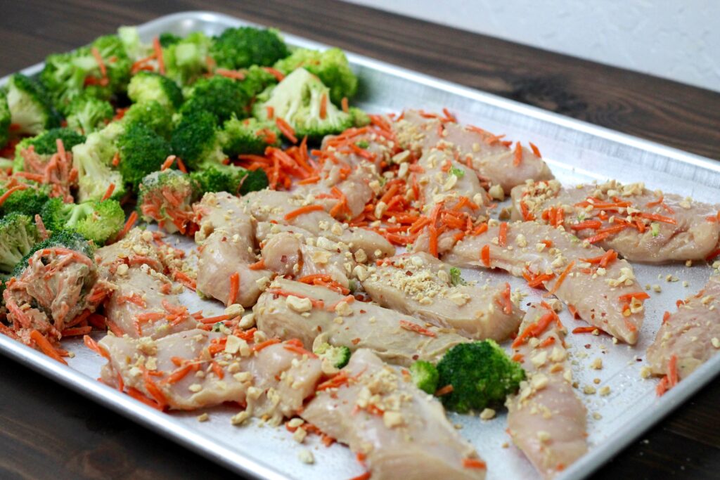 uncooked chicken topped with peanuts on one pan with a side of broccoli and shredded carrots